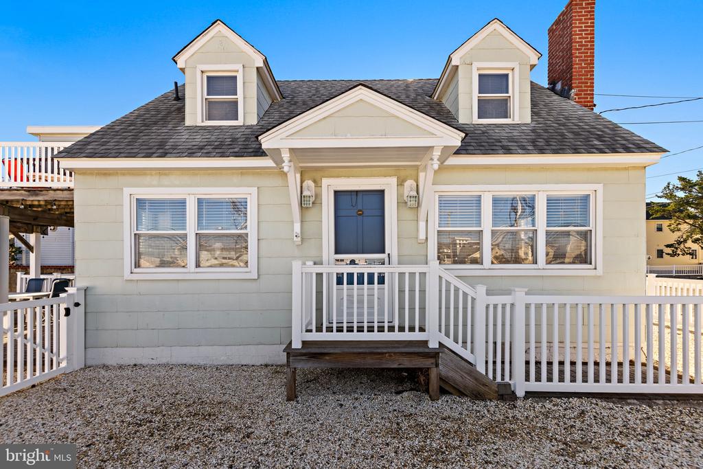 Light-colored house with two dormers, a blue front door, white railings, and a gravel yard.