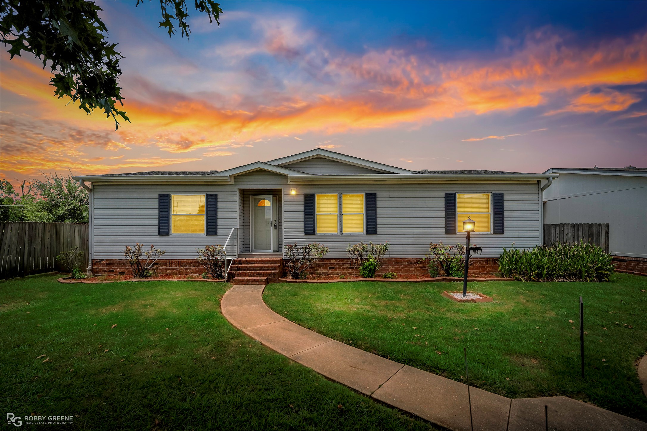 Single-story gray house with brick foundation and curved walkway beneath a colorful sunset sky.