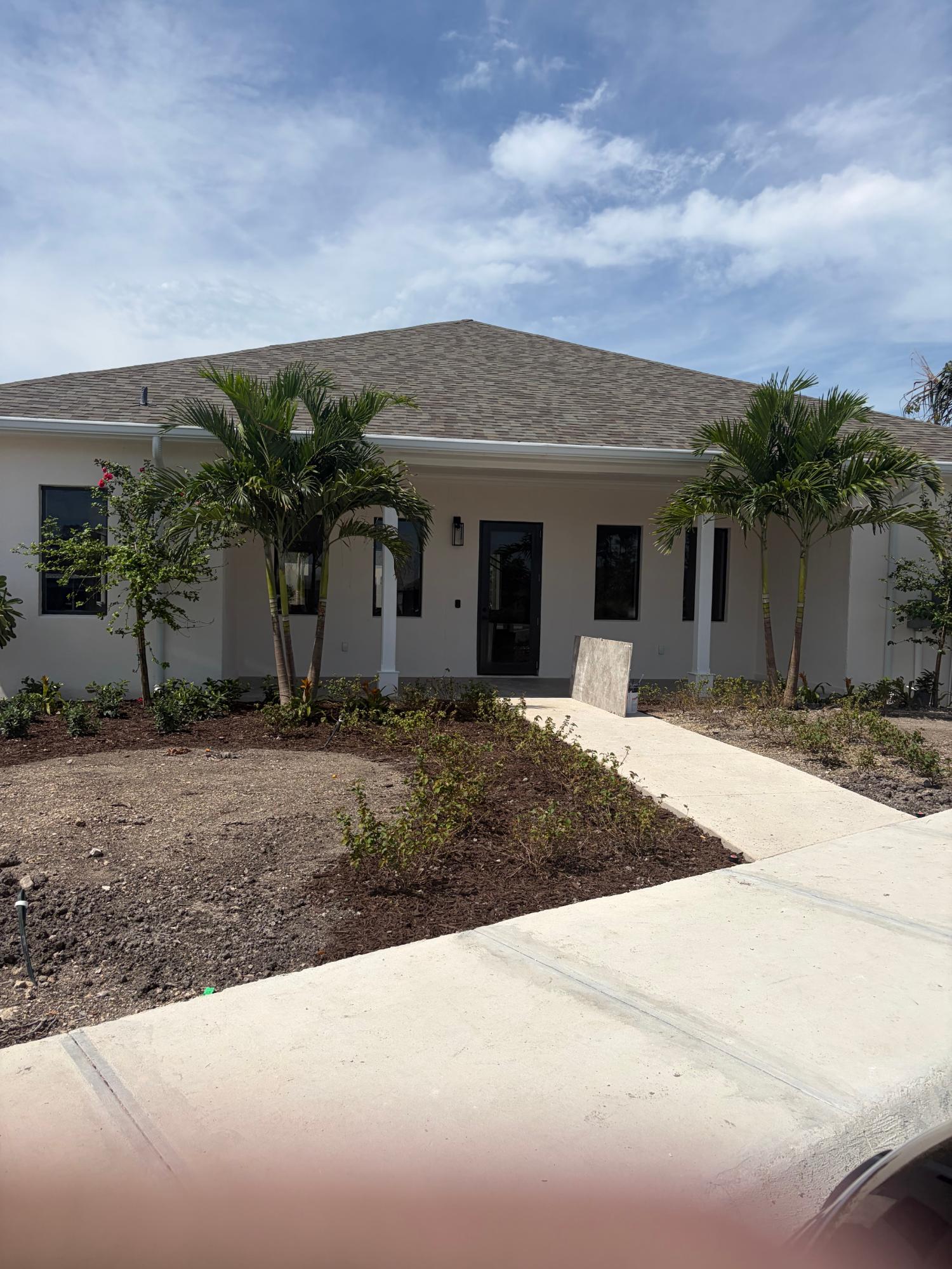 Single-story house with a broad shingled roof, palm trees, and a concrete walkway to the front entrance.