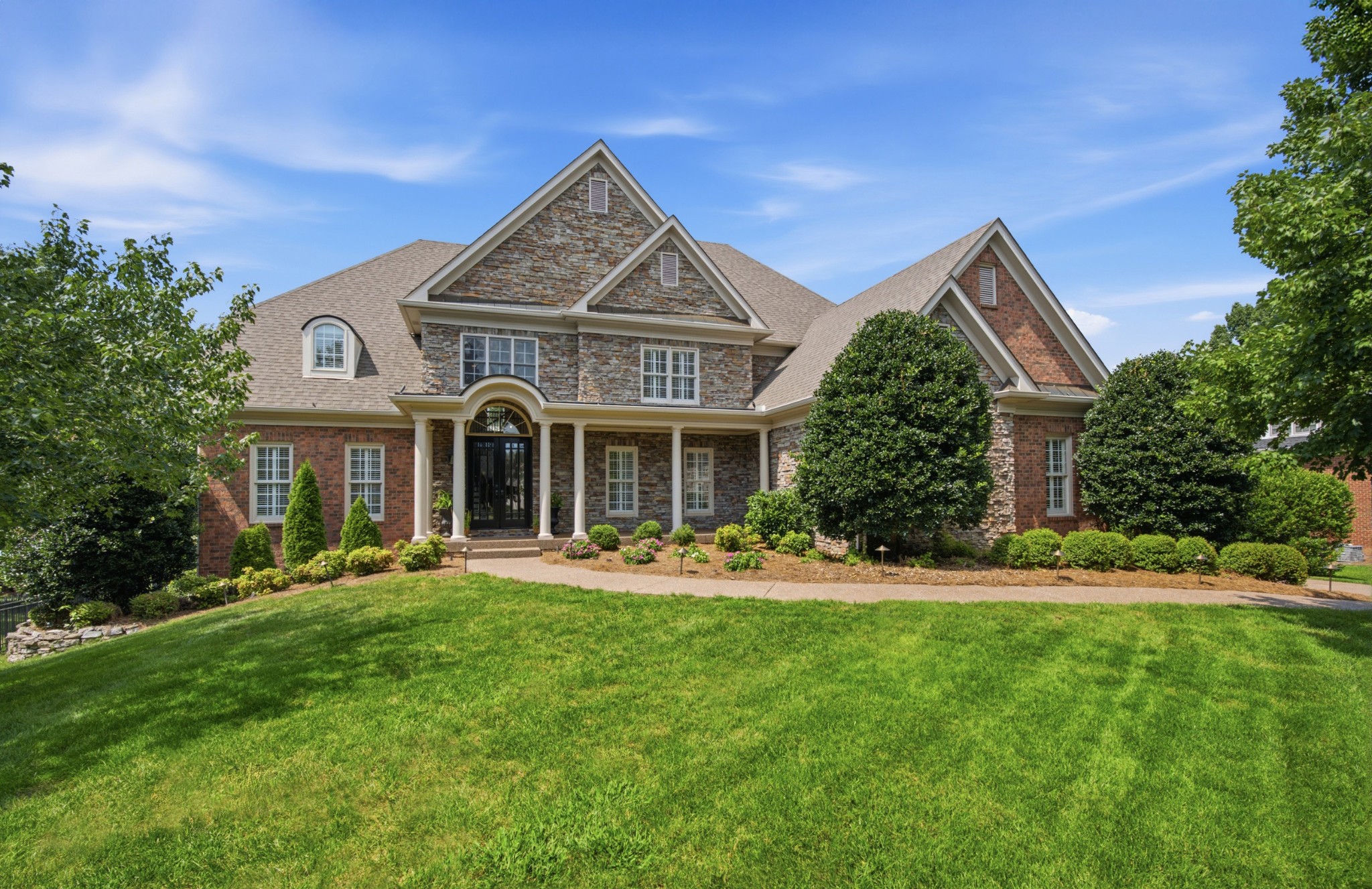 Two-story stone and brick home with a covered entry, landscaped shrubs, and a broad green lawn.