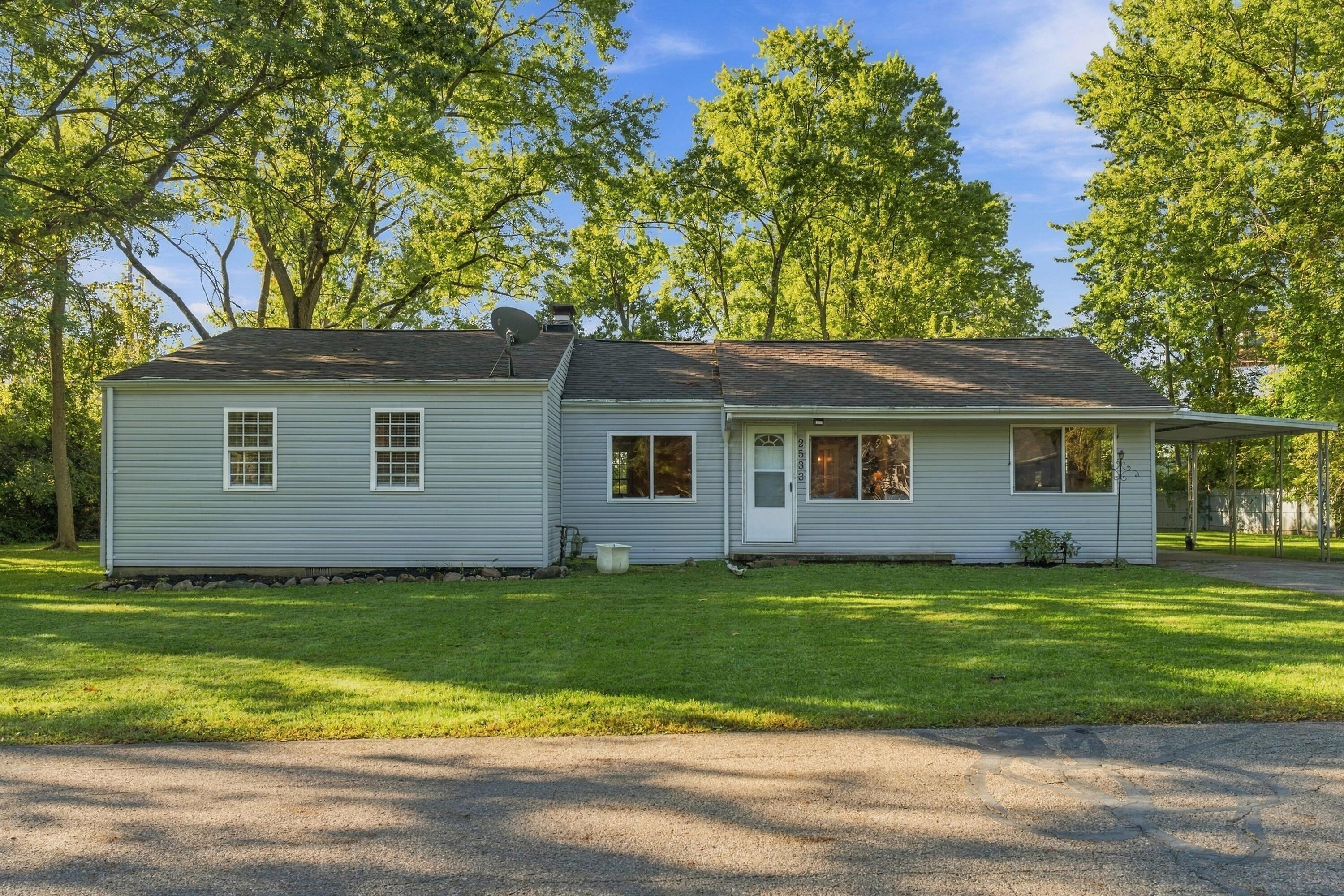 Single-story blue-gray home with a covered carport, broad lawn, and mature trees.