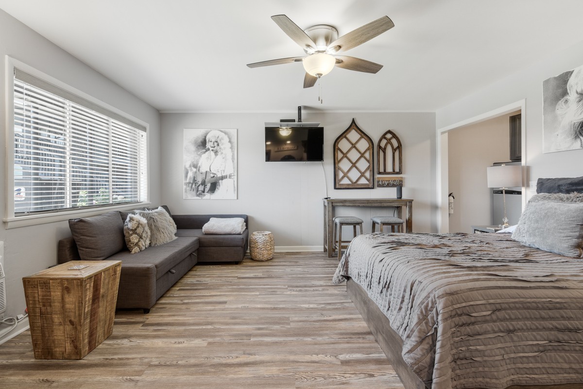 Bedroom with a gray sectional sofa, wide window with blinds, and a bed in the foreground.