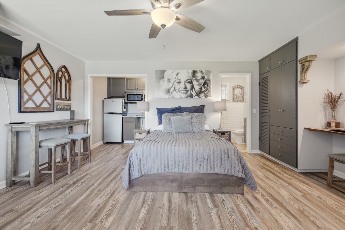 Furnished bedroom with a centered bed, ceiling fan, dark built-in cabinetry, and kitchenette beyond.