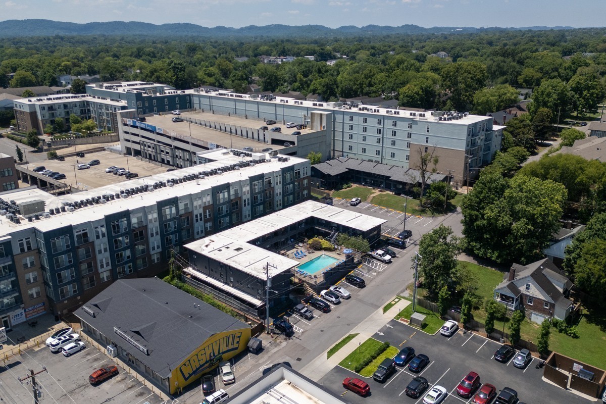 Aerial view of a modern apartment complex with a central pool and wooded hills beyond.