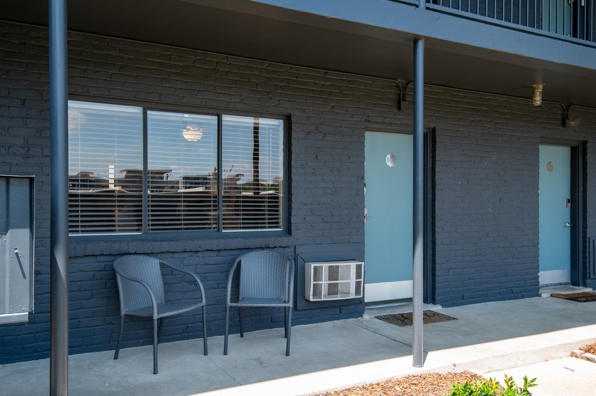 Apartment exterior with dark brick, blue unit 106 door, two patio chairs, and window air conditioner.