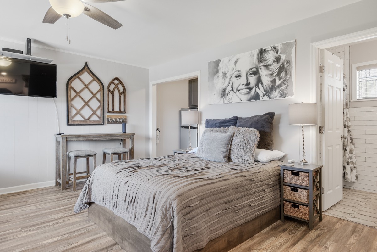 Furnished bedroom with gray bedding, a ceiling fan, and a bathroom doorway.