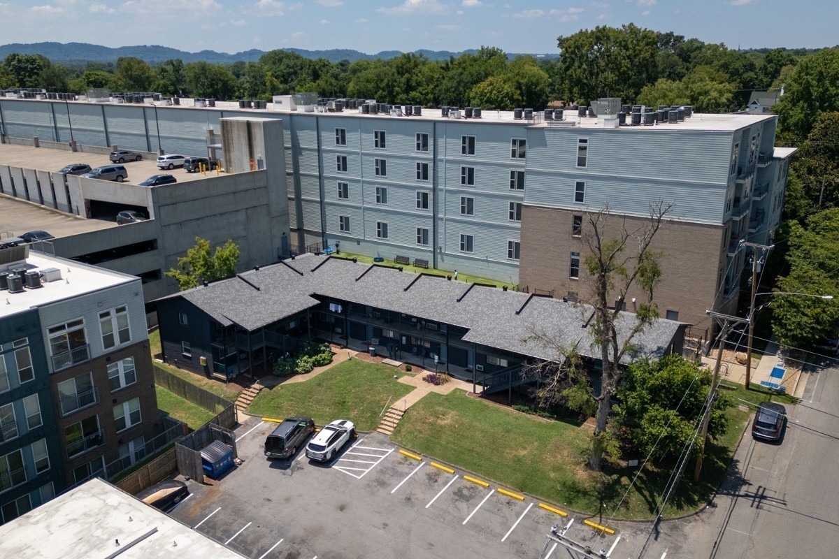 Aerial view of an apartment complex with a courtyard lawn, parking, and wooded hills beyond.