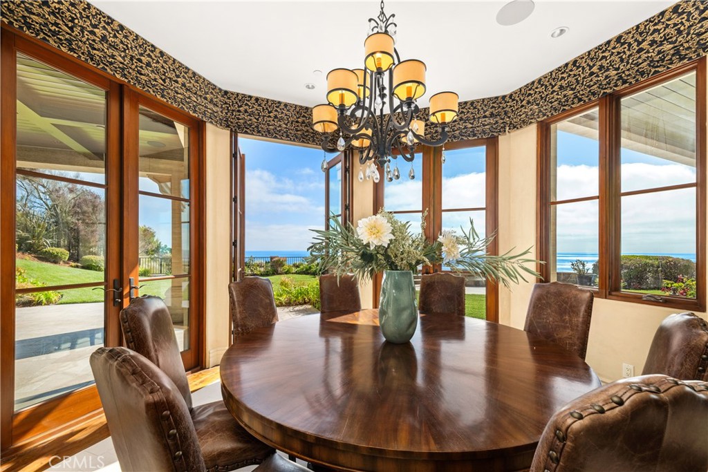 Dining room with an oval wooden table, chandelier, and ocean views through tall windows.