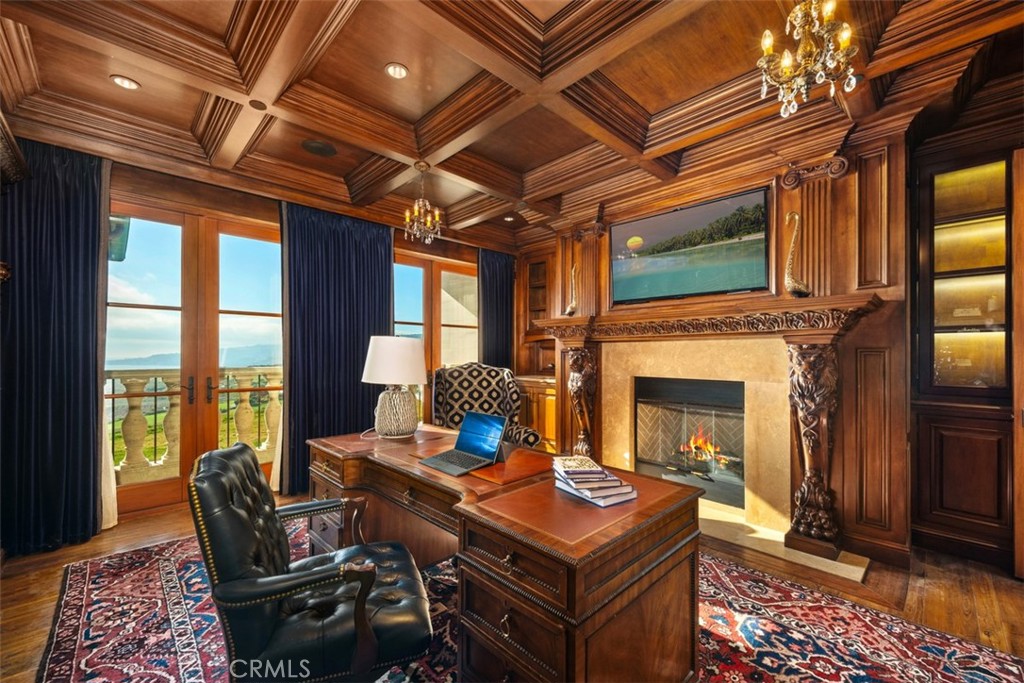 Wood-paneled home office with a carved fireplace, large desk, and ocean view through French doors.