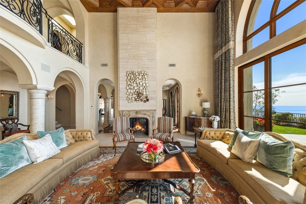 Living room with a stone fireplace, coffered ceiling, and ocean views through tall arched windows.