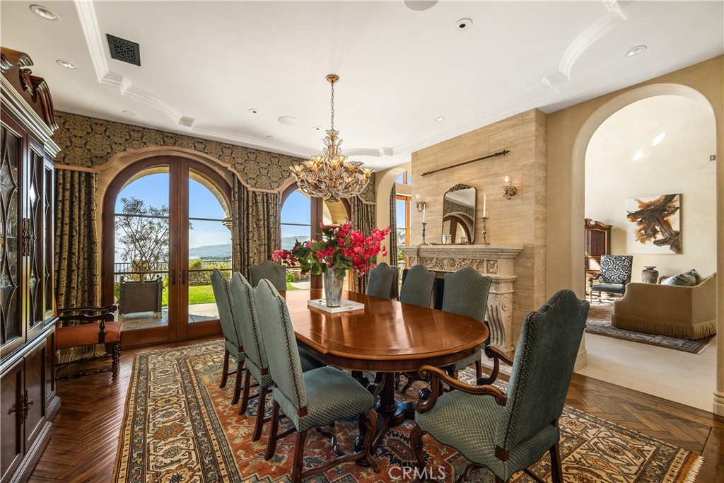 Furnished dining room with an oval table, ornate chandelier, arched glass doors, and carved stone fireplace.