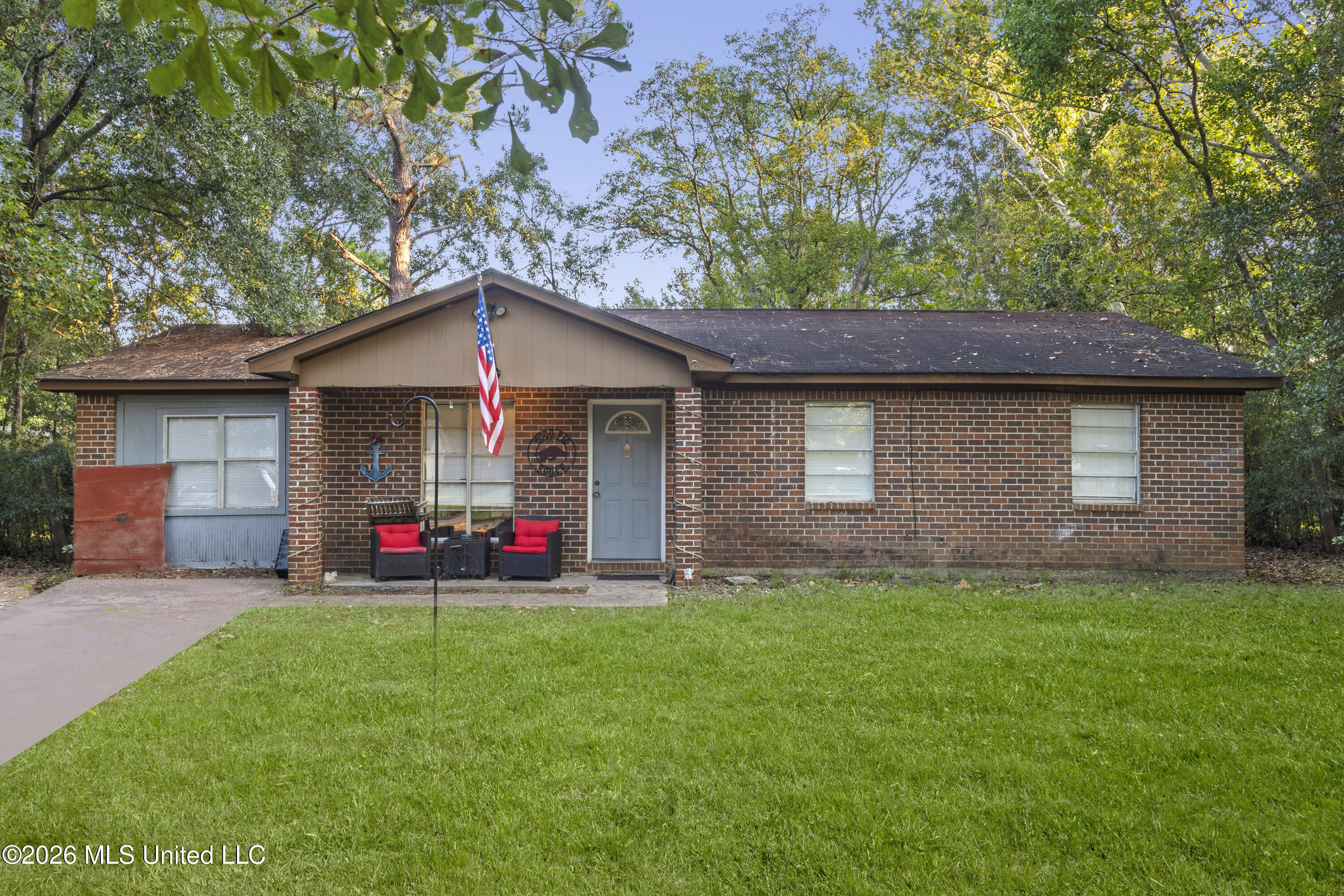 Single-story brick house with a blue-gray door, American flag, red-cushioned porch chairs, and wooded lawn.