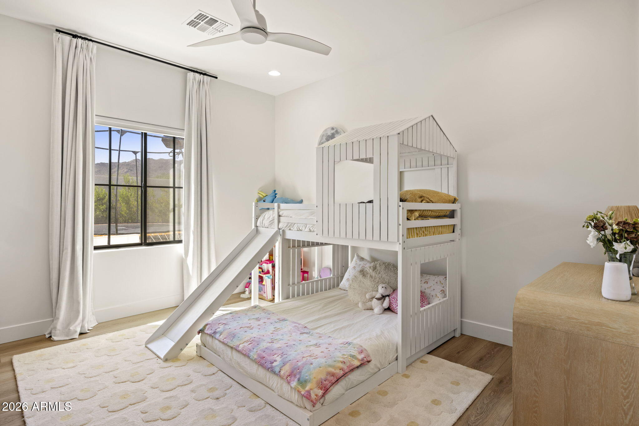 Children’s bedroom with a white house-style bunk bed and slide beside a large window.