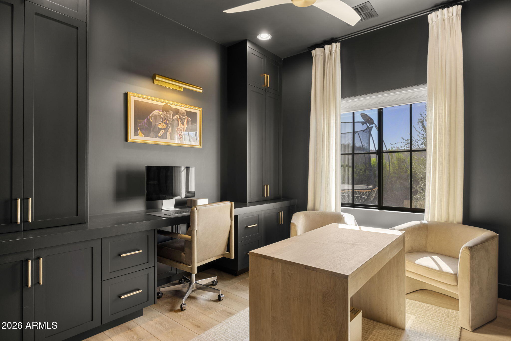 Home office with dark built-ins, a light wood table, cream chairs, and a large curtained window.