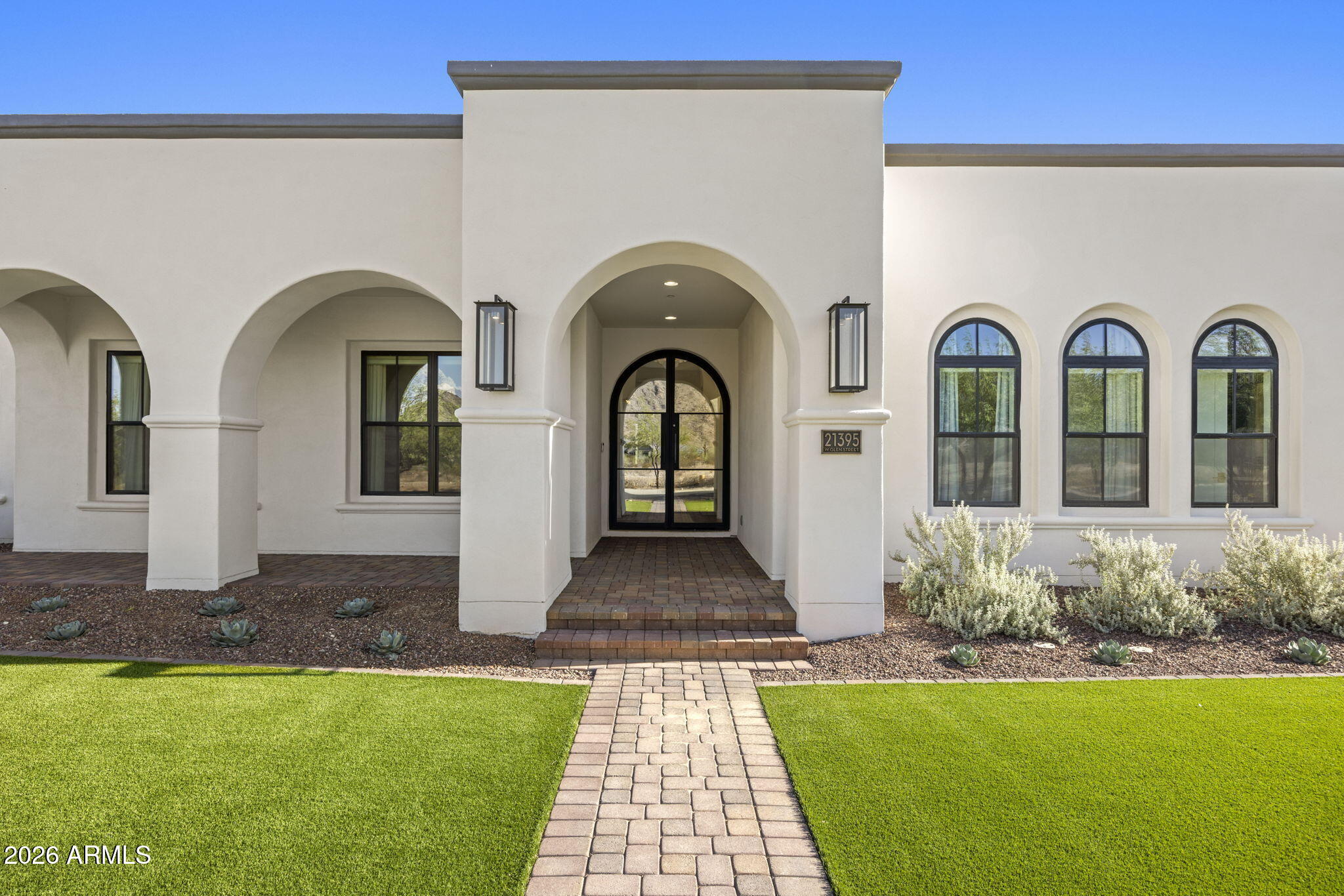 White stucco home with an arched entry, black-framed windows, and a brick walkway across the lawn.