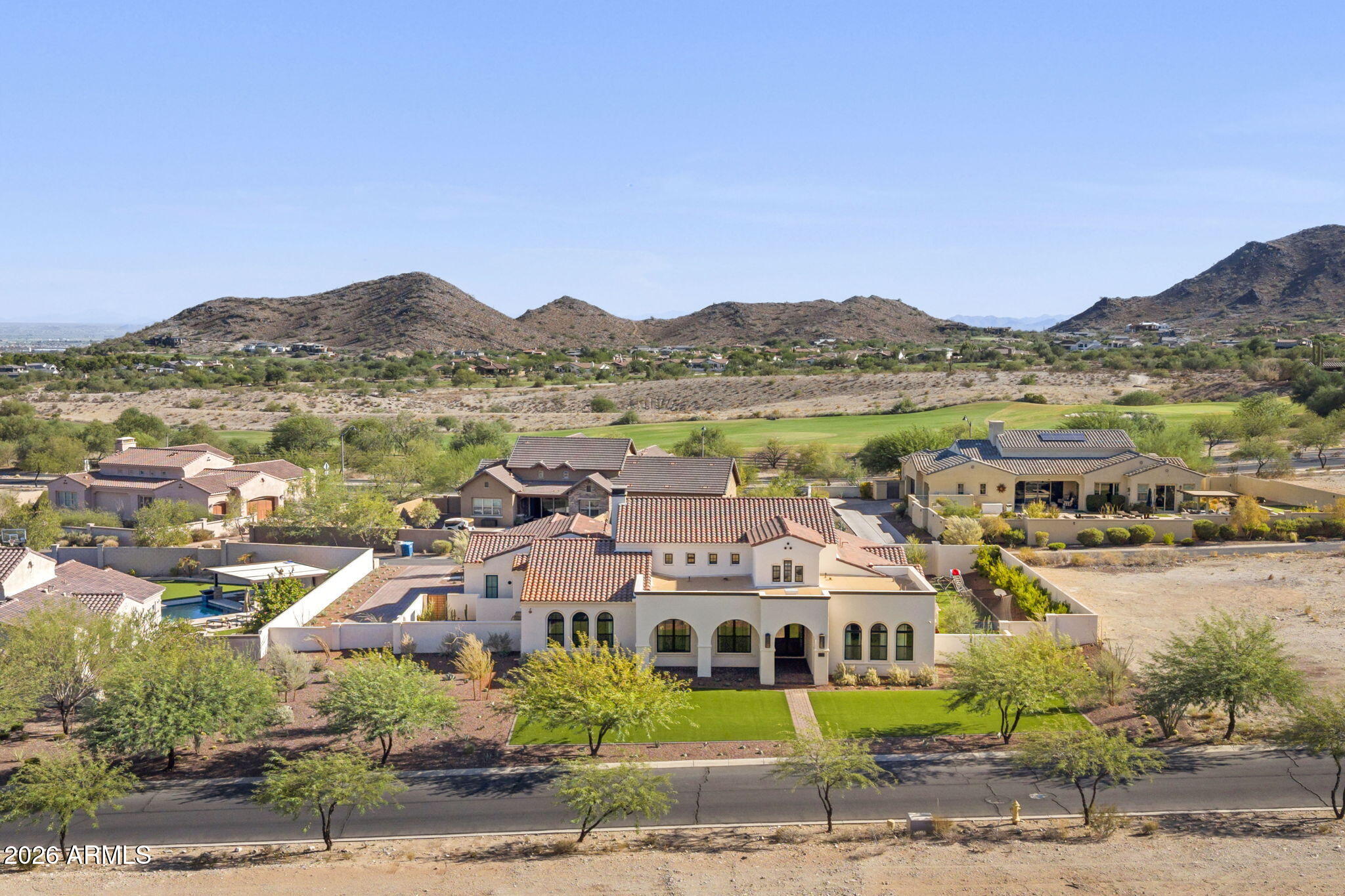 Large stucco home with a red tile roof, green front lawn, golf course, and rocky hills beyond.