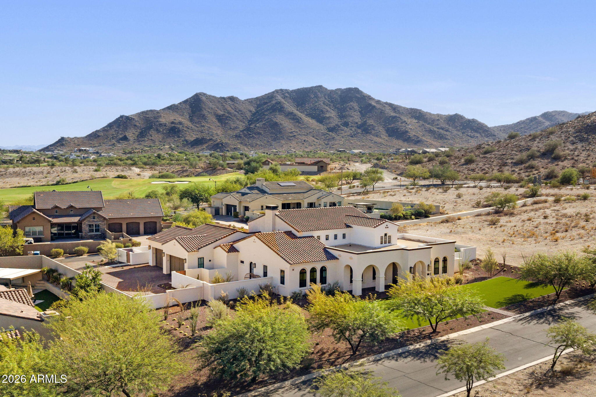 Aerial view of a white stucco home with an arched patio, green lawn, golf course, and desert mountains.