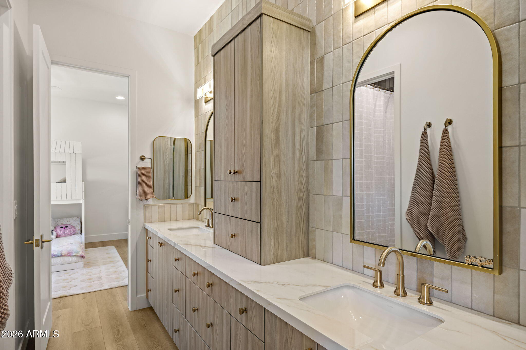 Bathroom with a long double vanity, brass fixtures, and large arched mirrors beside an open bedroom doorway.
