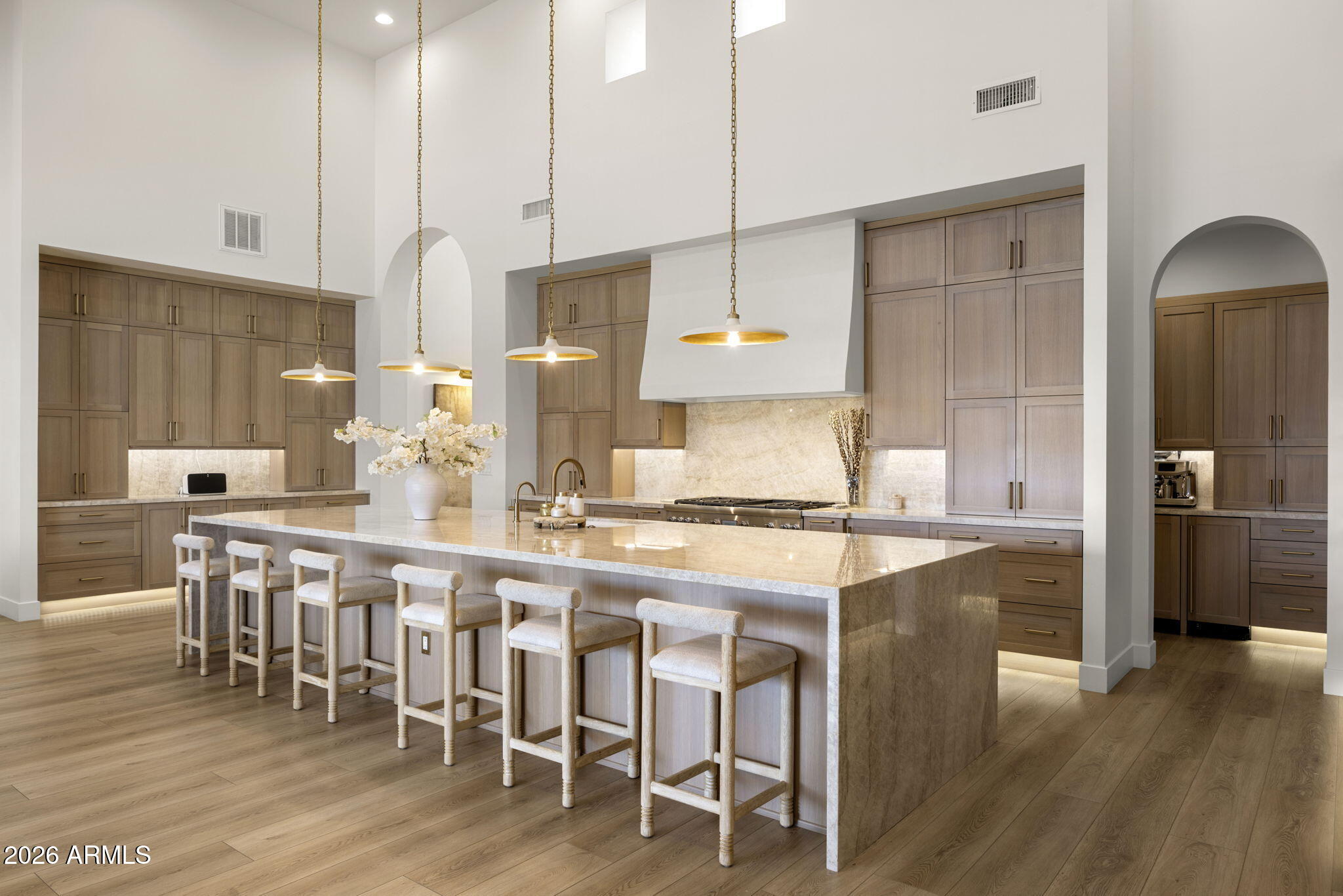 Kitchen with a large marble island, six upholstered stools, tall wood cabinets, and brass pendant lights.