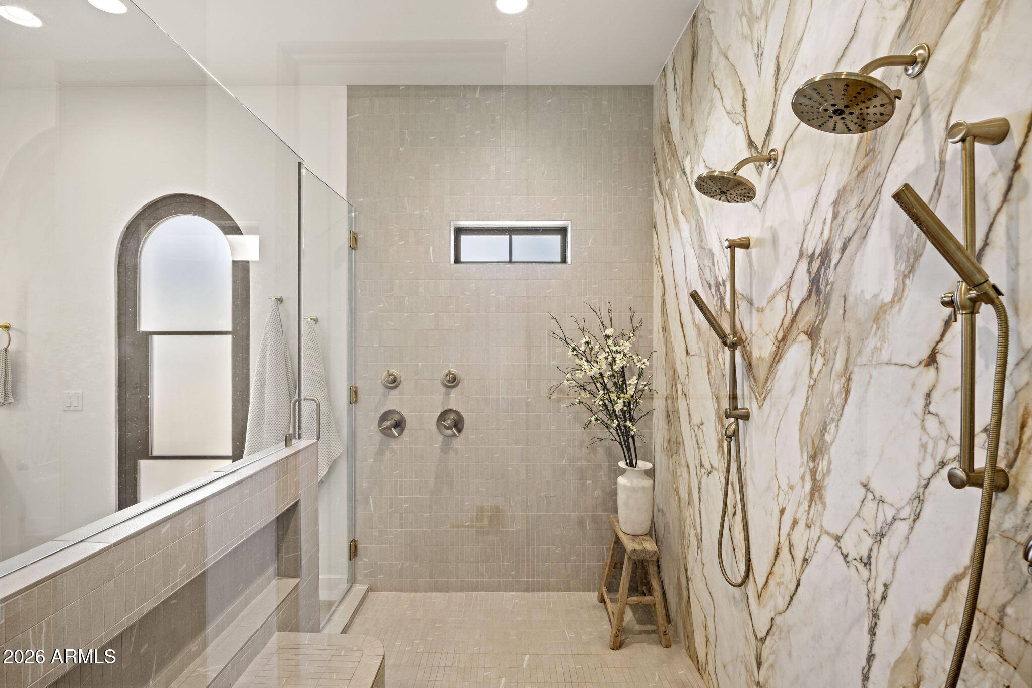 Walk-in shower with a veined marble accent wall, glass partition, and multiple brass showerheads.