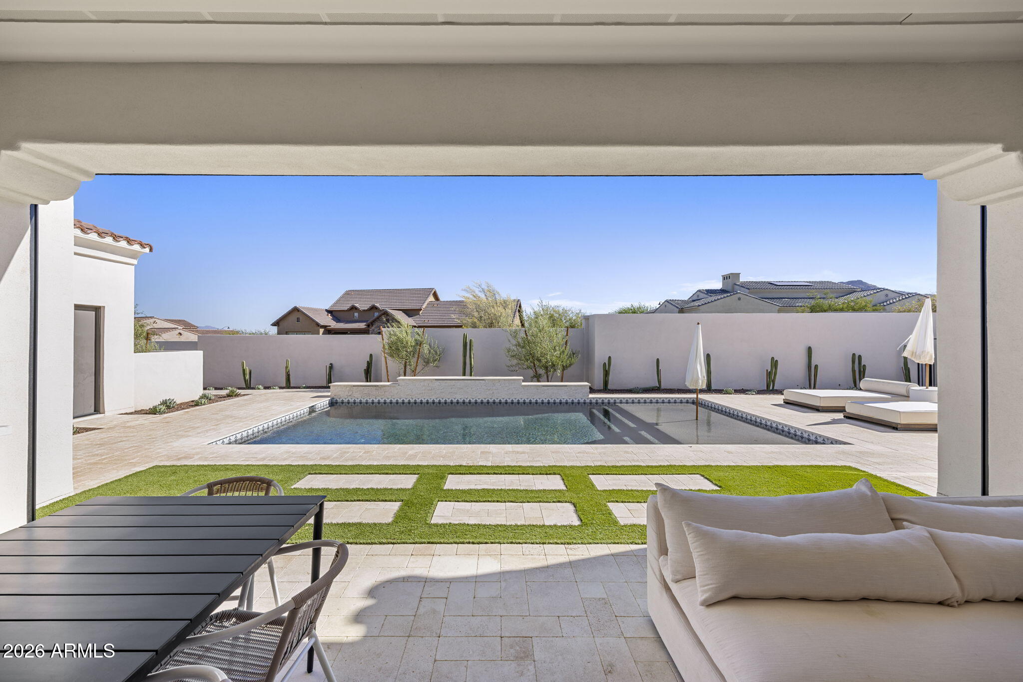 Backyard pool viewed from a covered patio, with lounge chairs and a strip of lawn.