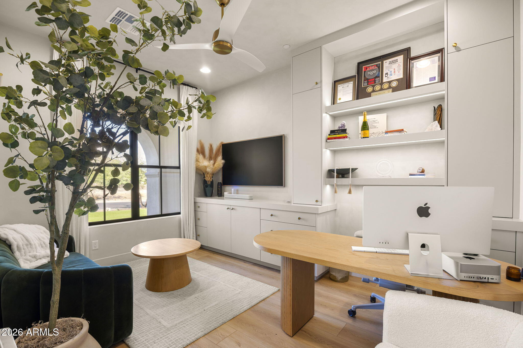 Furnished home office with built-in display shelves, a wooden desk, and a large window.