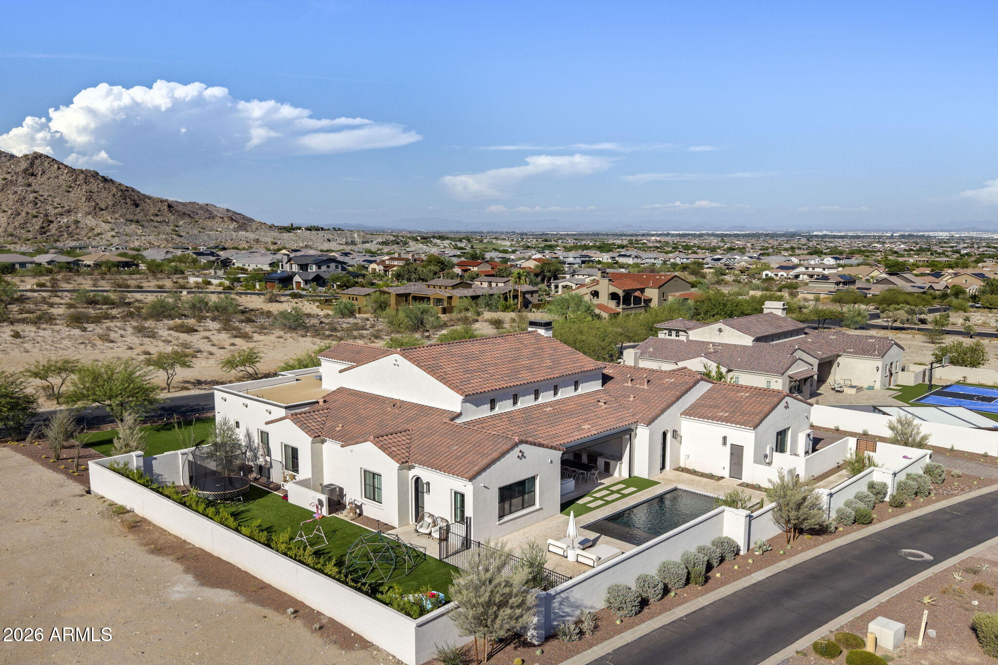 Aerial view of a white stucco home with a backyard pool, green lawn, and rocky hillside beyond.