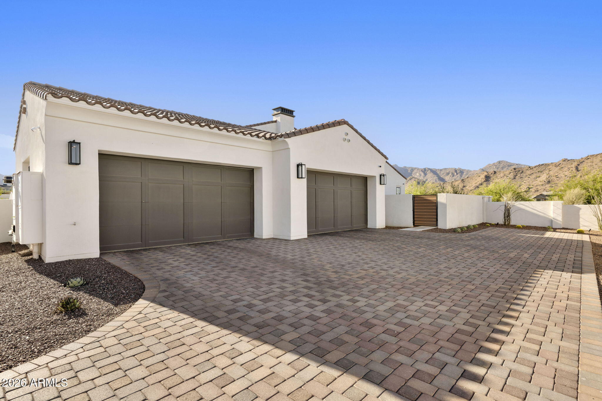 Home exterior with two dark garage doors, a wide paver driveway, and rocky mountains beyond the wall.