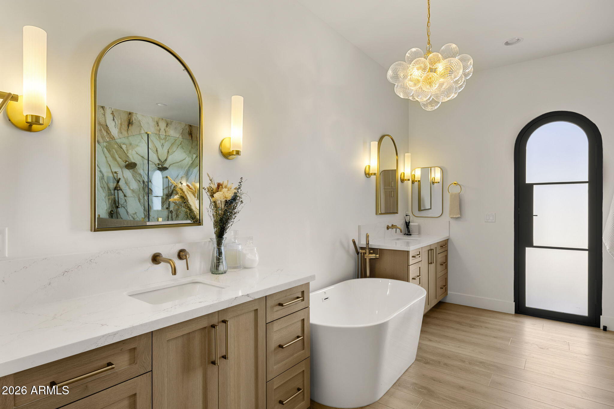 Primary bathroom with double vanity, freestanding tub, brass fixtures, and an arched black-framed window.