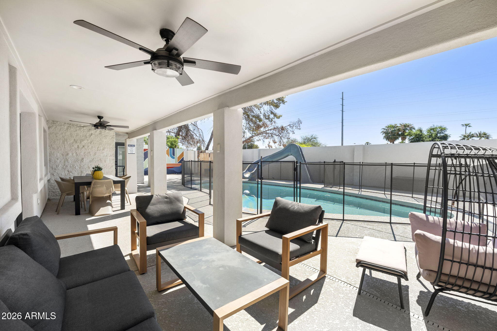 Covered patio with lounge seating beside a fenced pool and slide.