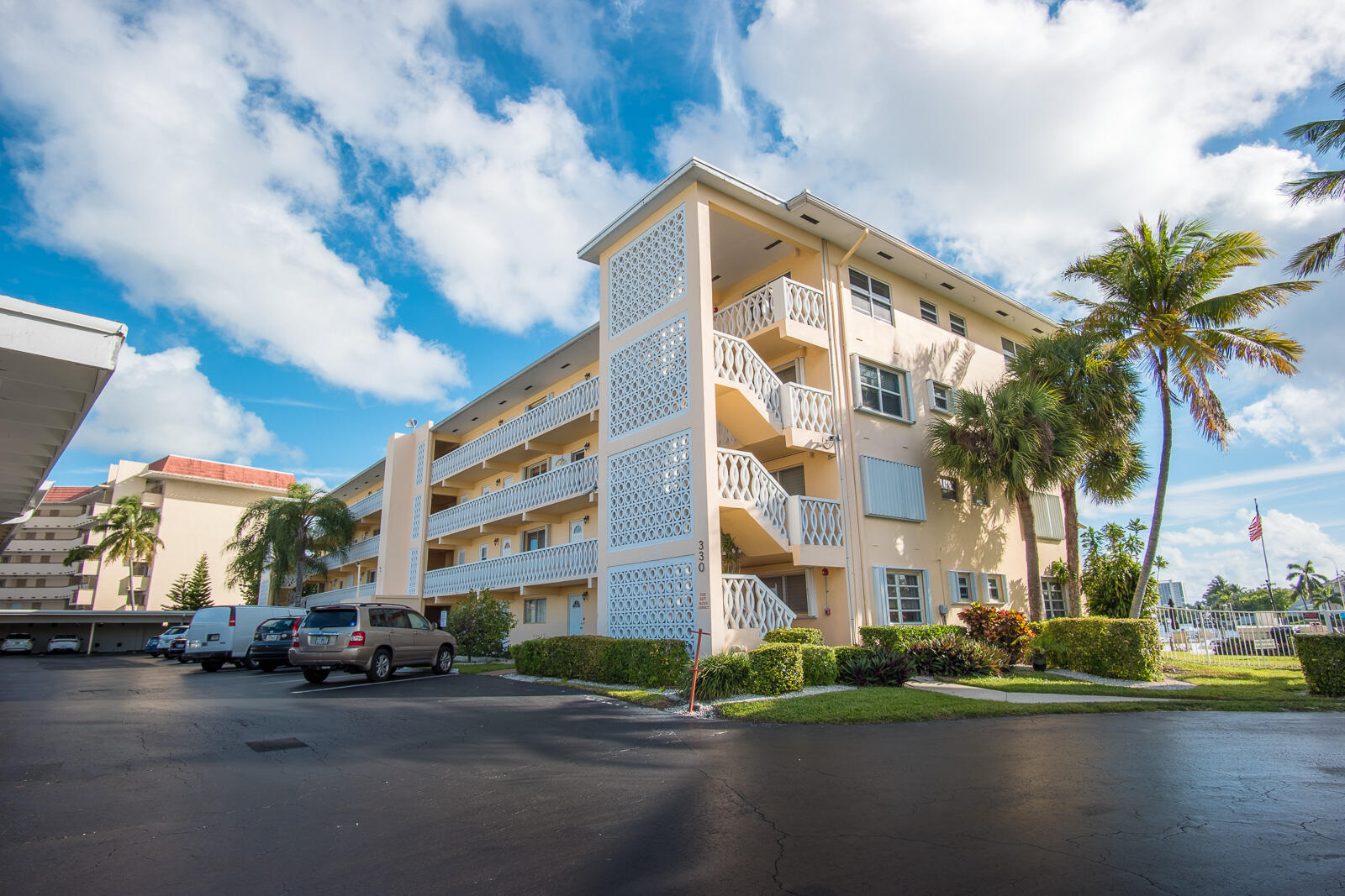 Low-rise condominium with white balcony railings, exterior stairs, and palm trees beside the parking lot.