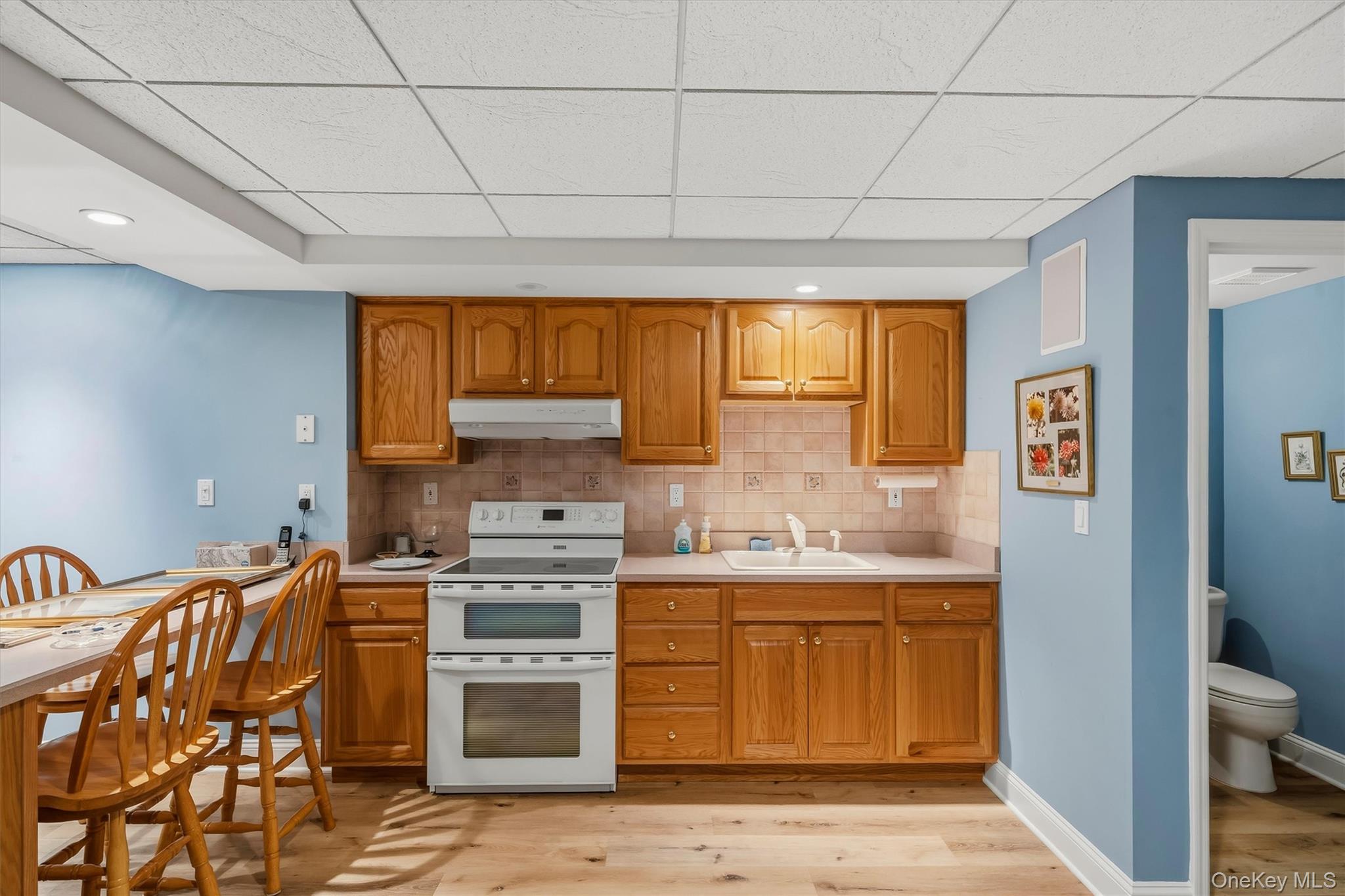 Kitchen with honey-colored cabinets, white appliances, and a dining table beside a bathroom doorway.