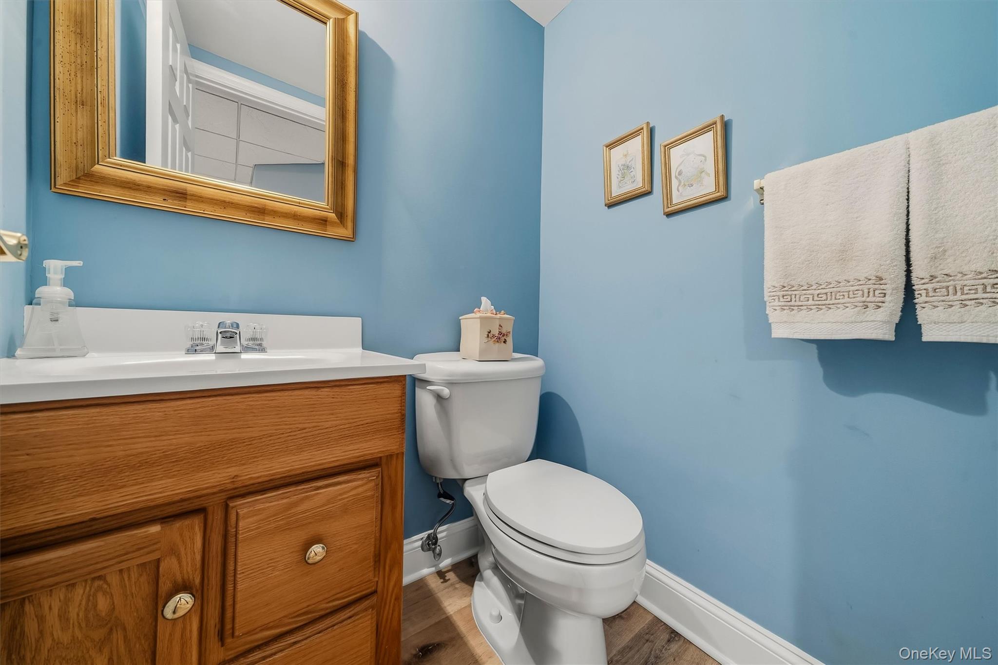 Bathroom with blue walls, a wood vanity, white sink and toilet, and gold-framed mirror.