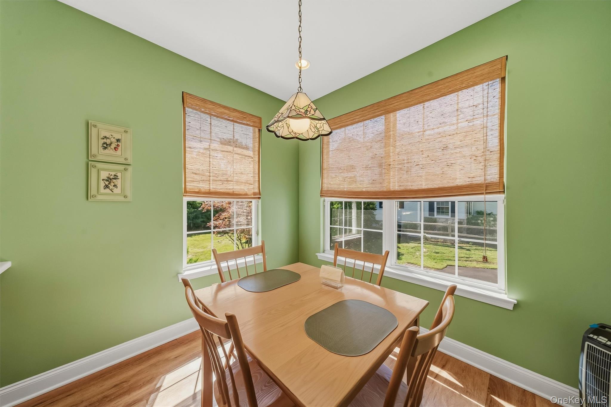 Dining room with sage-green walls, a wooden table, bamboo shades, and two large windows.
