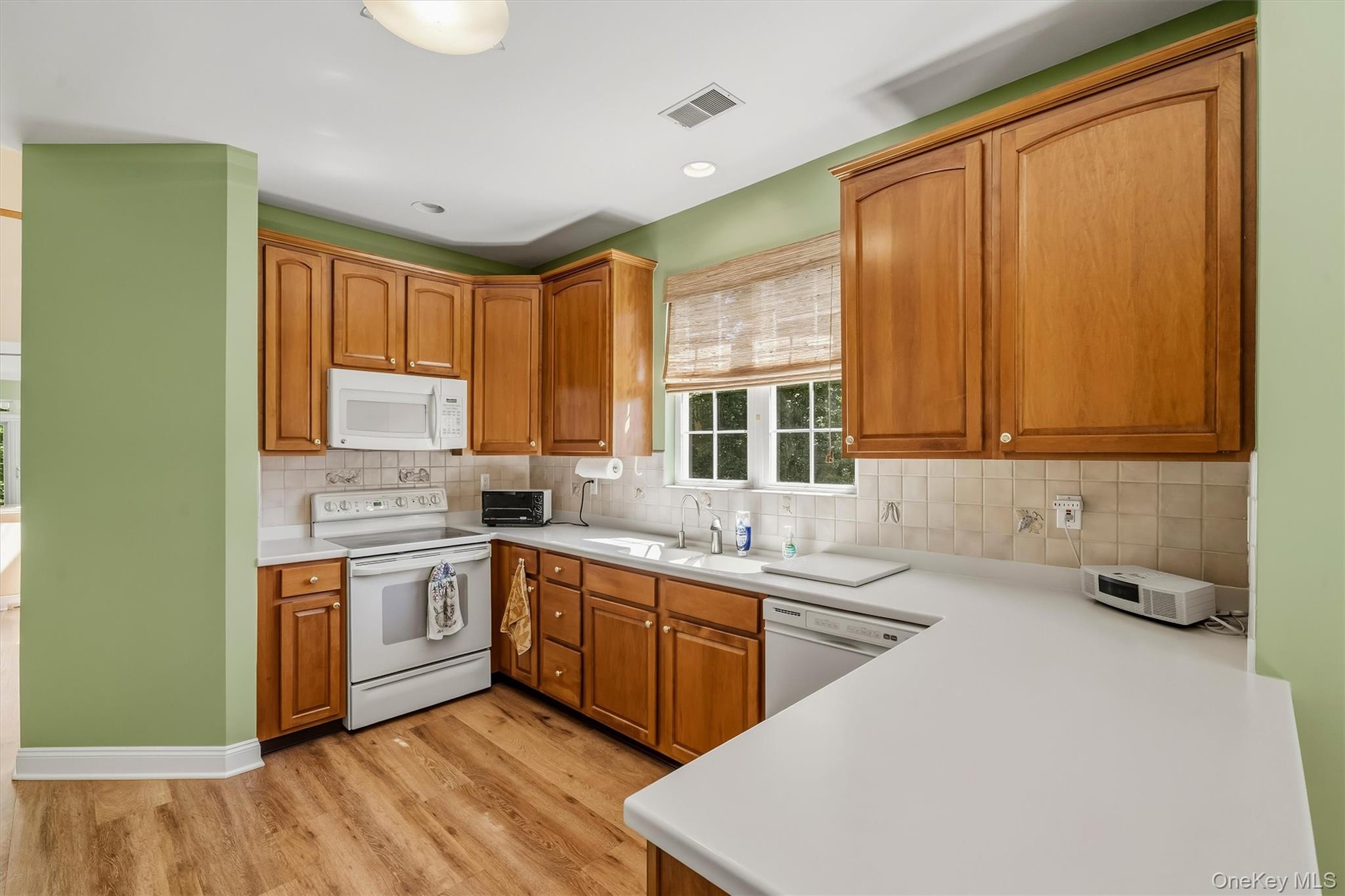 Kitchen with honey-toned wood cabinets, white countertops, green walls, and a sunlit window.