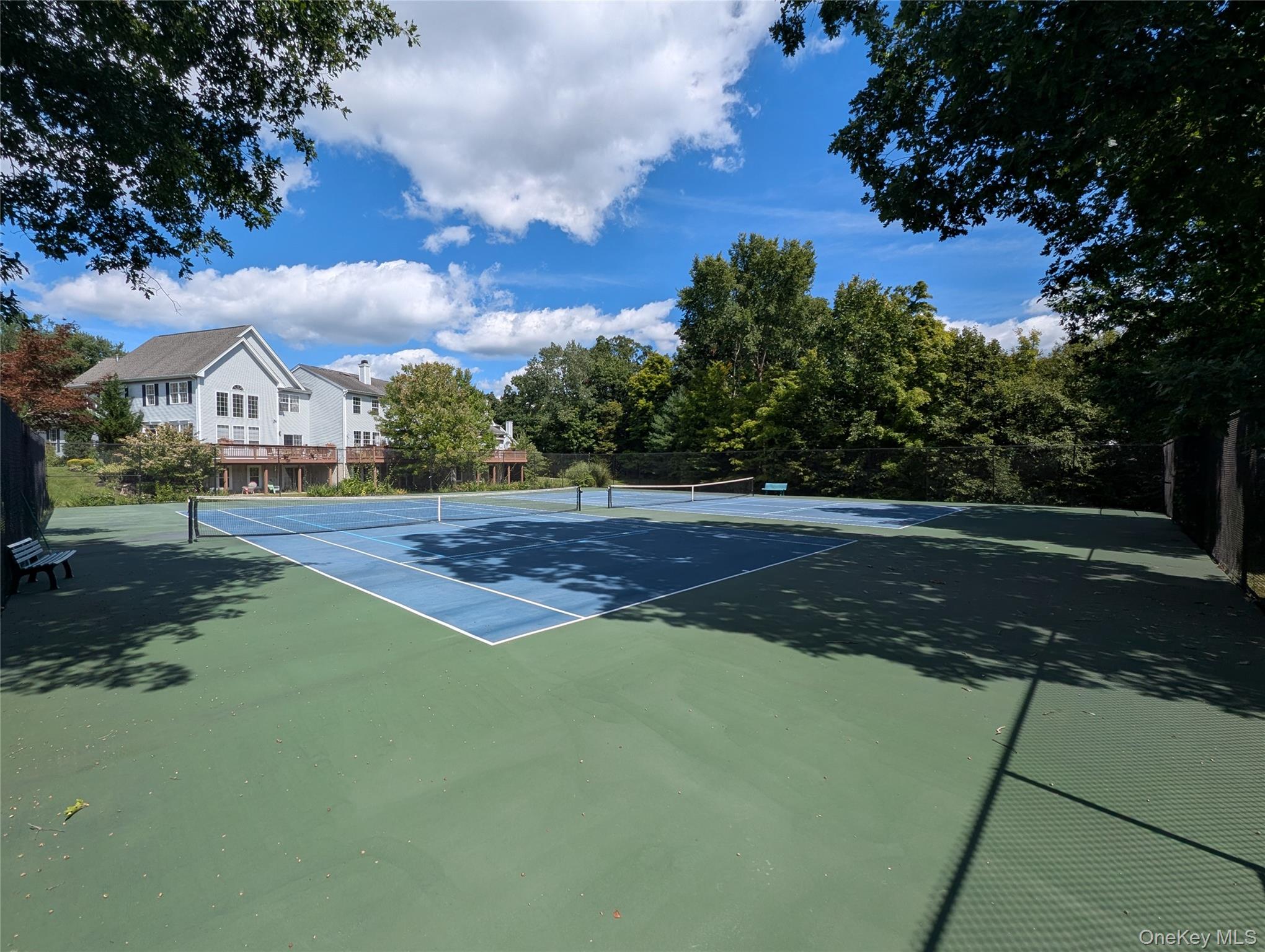Fenced tennis court beside a large house, shaded by mature trees.