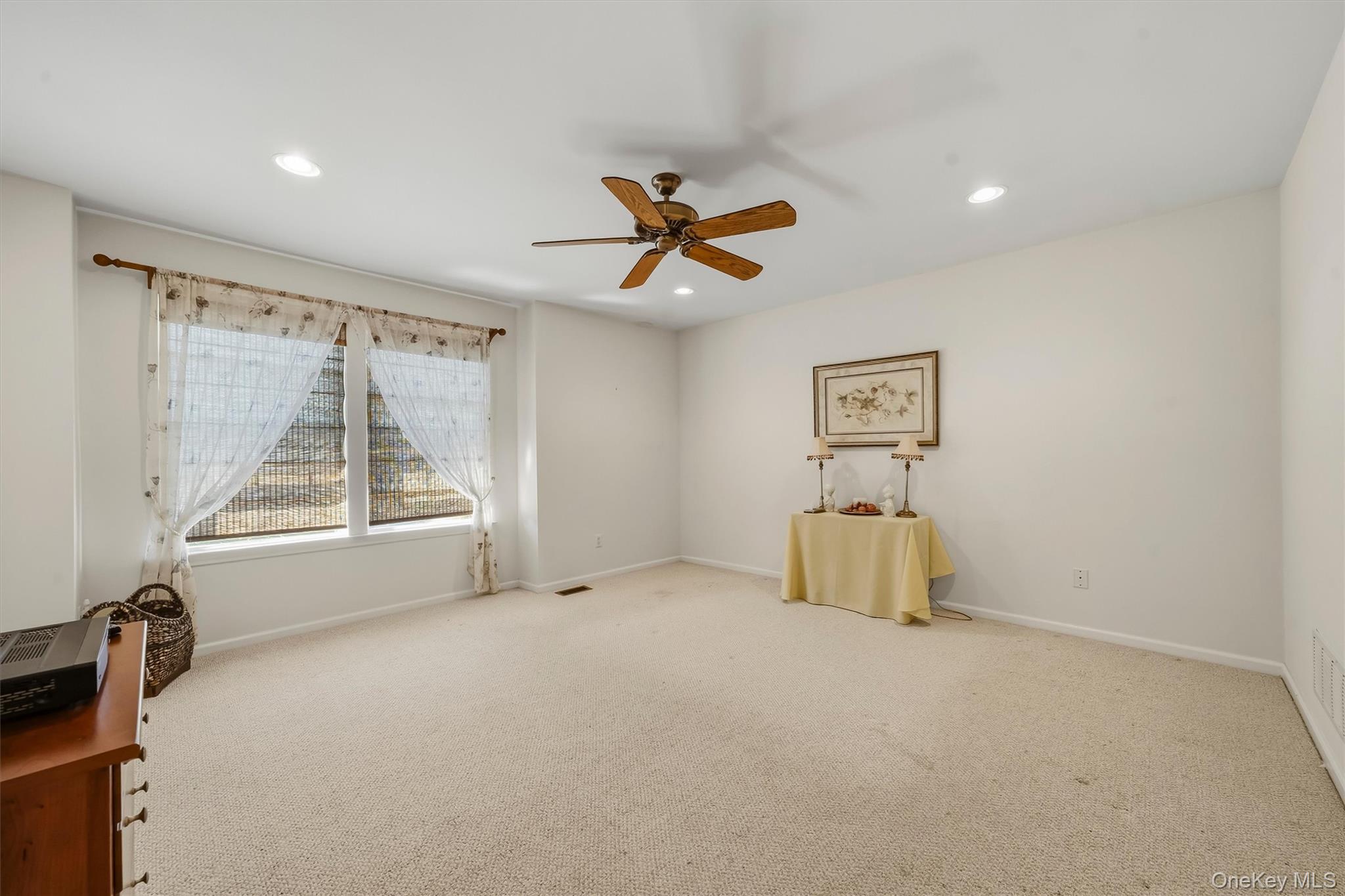 Carpeted room with a large curtained window, wood-blade ceiling fan, and small table beneath framed artwork.