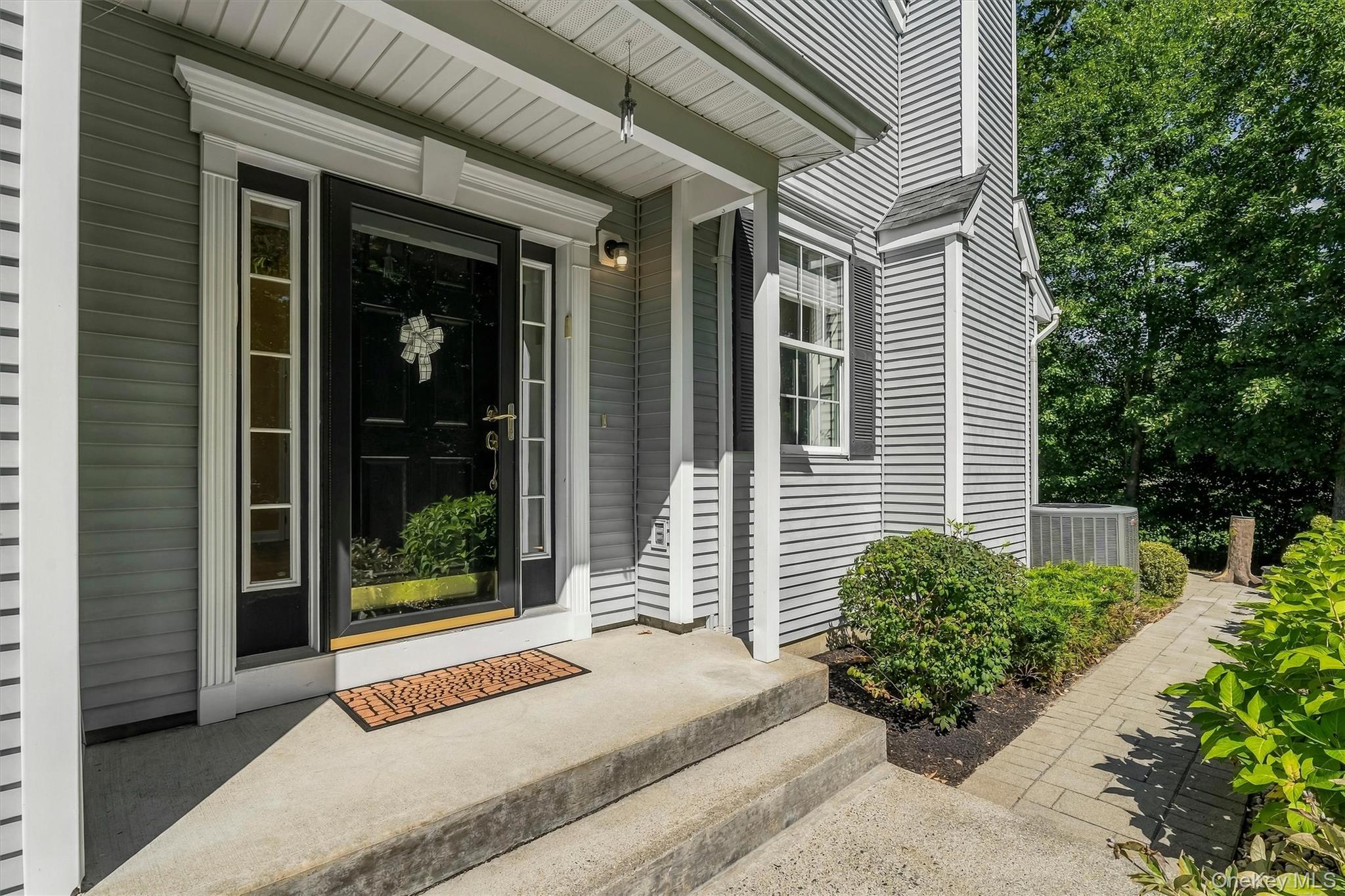 Gray-sided home entrance with a black door, covered porch, concrete steps, and shrub-lined walkway.
