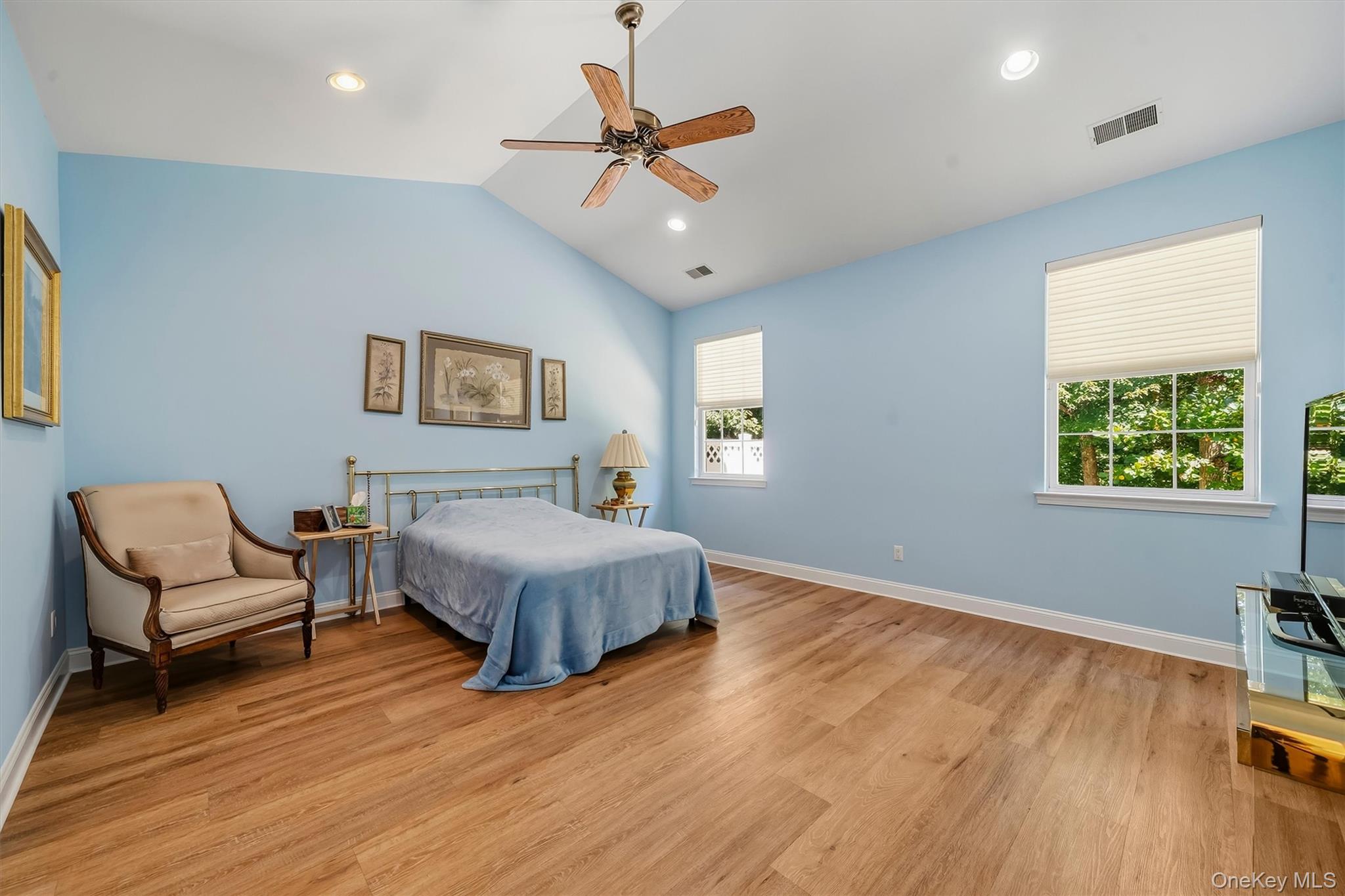Bedroom with pale blue walls, hardwood floors, a blue-covered bed, and windows overlooking greenery.
