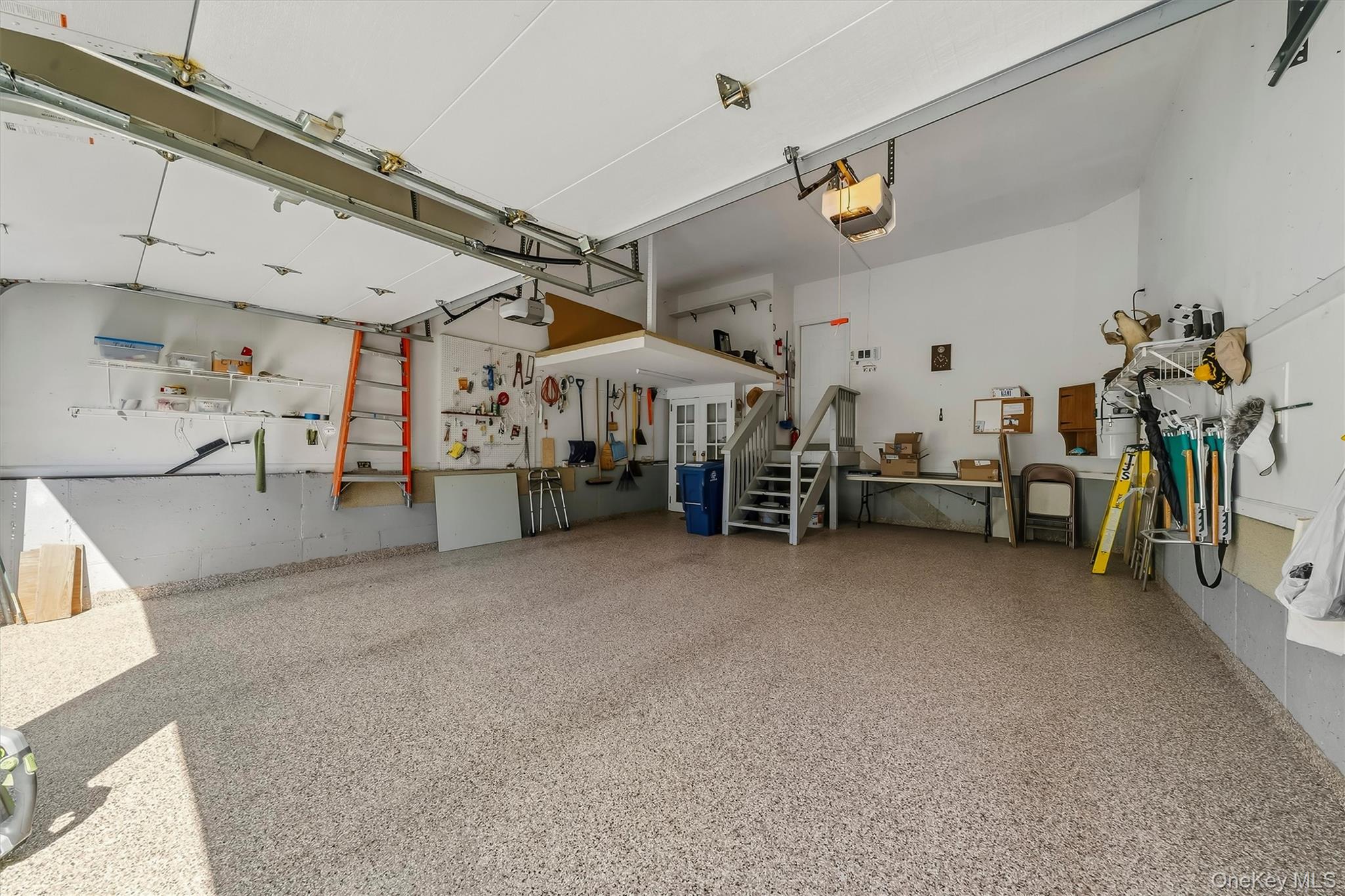 Spacious garage with a speckled floor, wall-mounted tools, workbench, and stairs to an upper level.