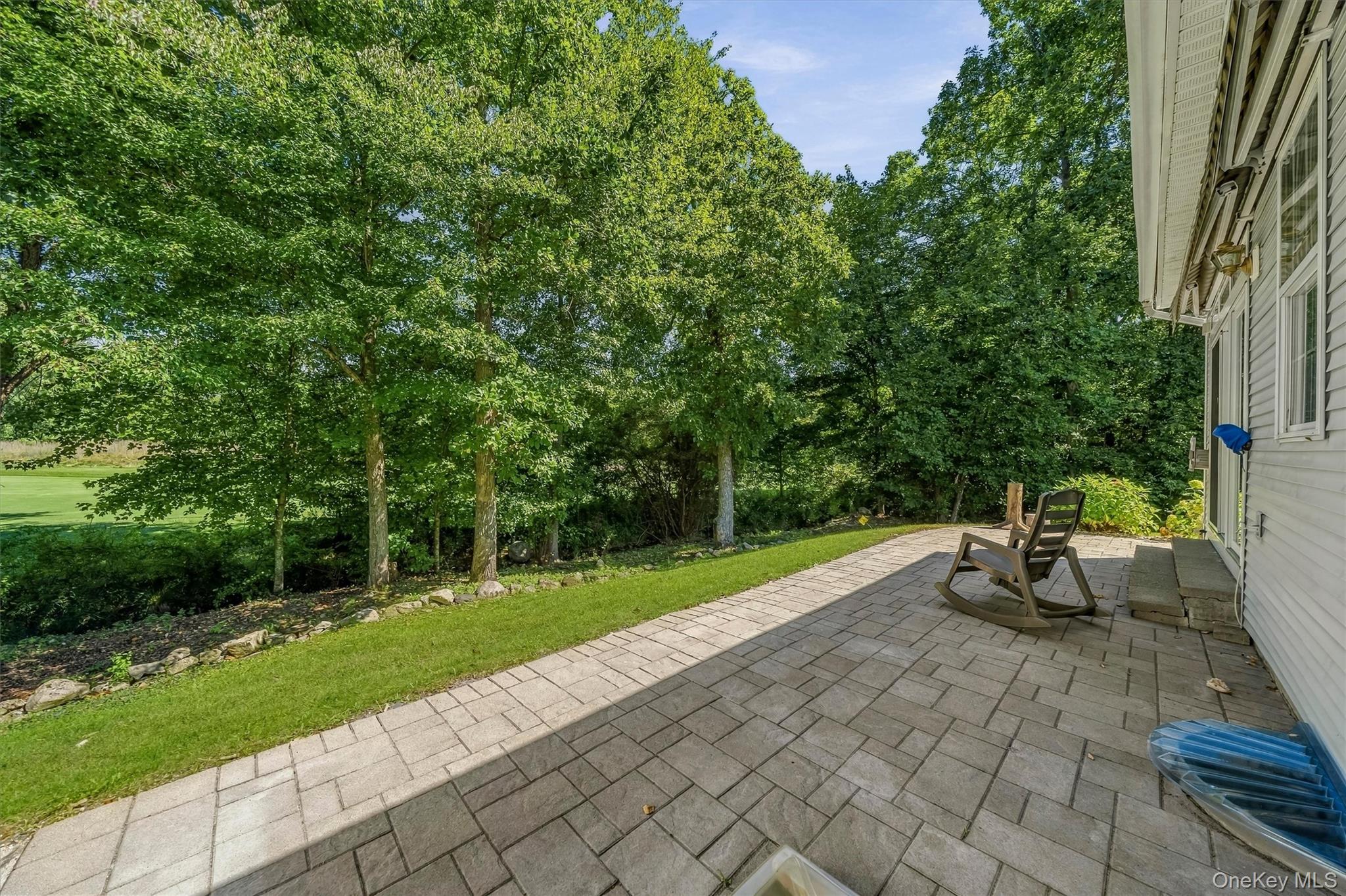 Paver patio beside the house with rocking chairs, a strip of lawn, and mature trees.
