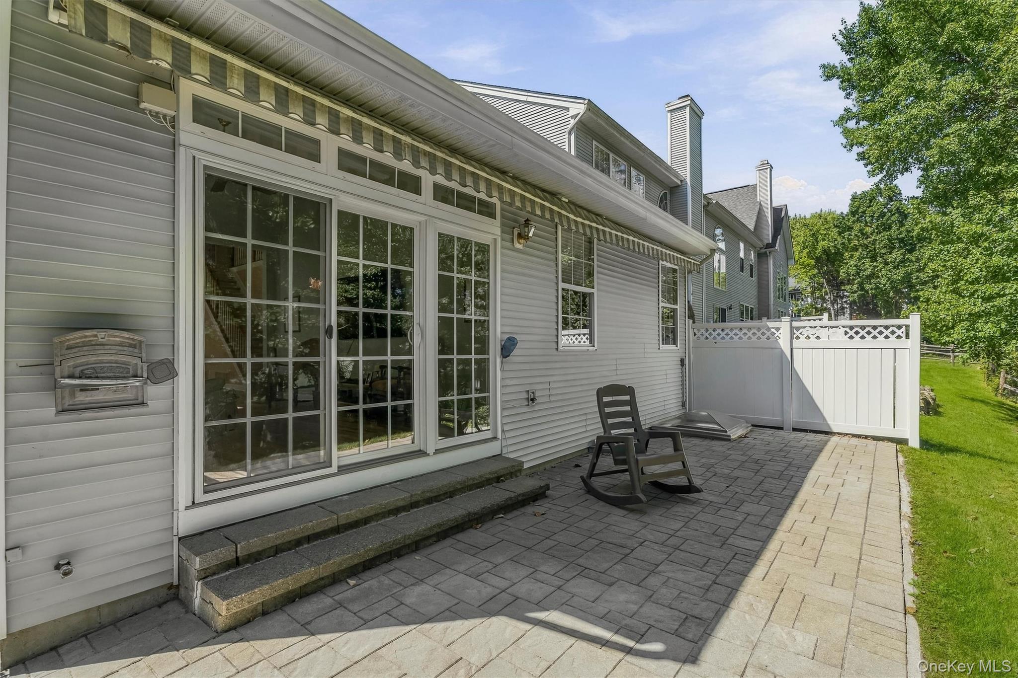 Paved patio beside a gray-sided home, with sliding doors, a striped awning, and a white privacy fence.