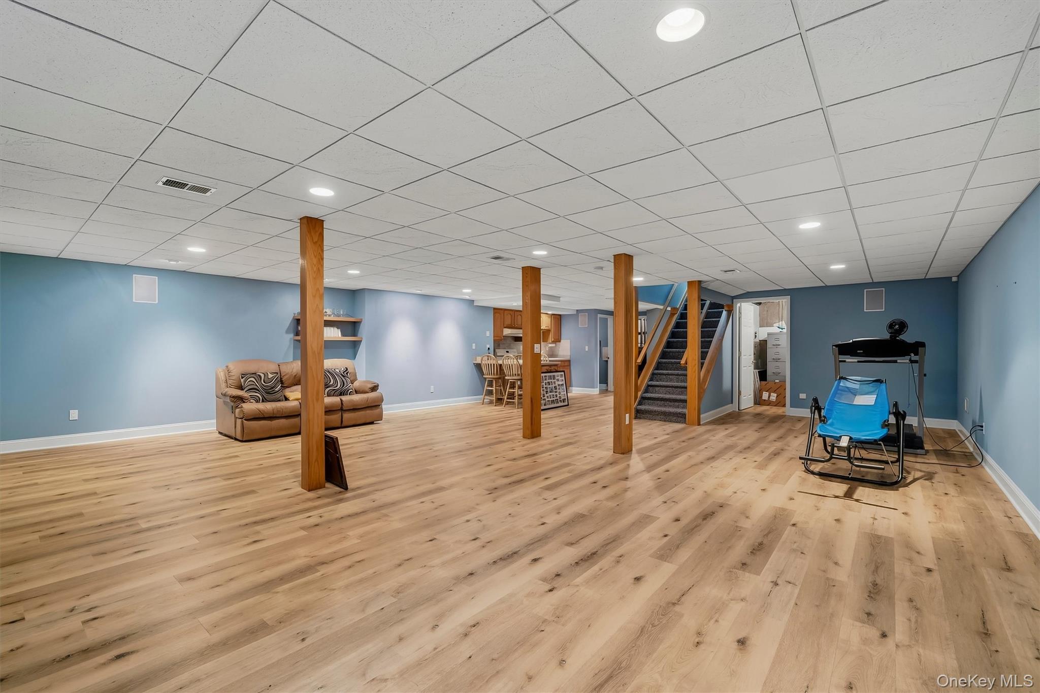 Finished basement recreation room with wood floors, blue walls, support posts, sofa, exercise equipment, and stairs.