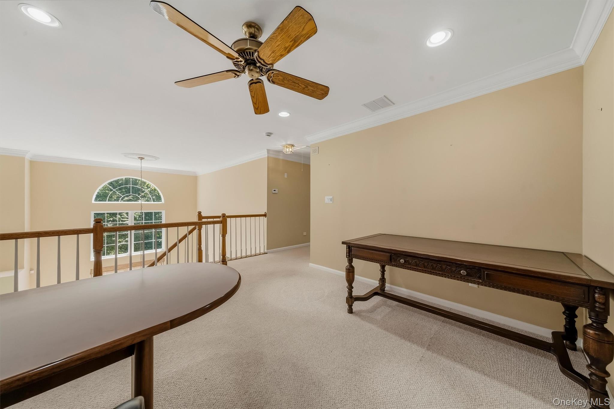 Upstairs carpeted landing with an arched window, open railing, and two dark wood tables.