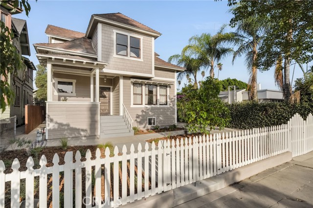 Two-story beige house with a covered porch behind a white picket fence and palm trees.