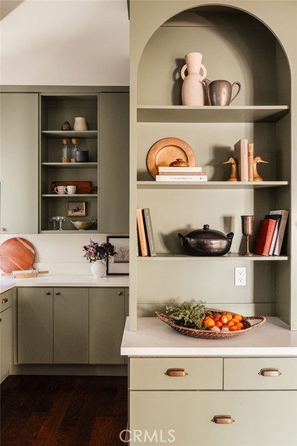 Sage-green kitchen with arched display shelves, pottery, cookbooks, and a bowl of oranges.