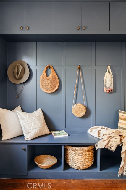 Blue built-in entryway bench with hanging bags, hats, pillows, and woven storage baskets.