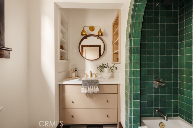 Bathroom vanity beside a dark green tiled shower, with a round mirror and brass lights.