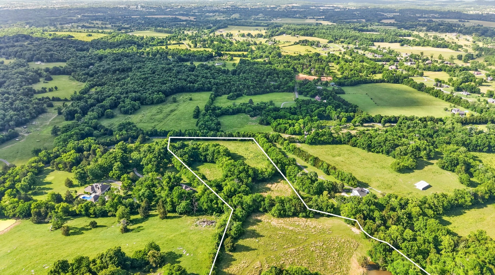 Aerial view of a wooded rural parcel outlined in white among fields and nearby homes.