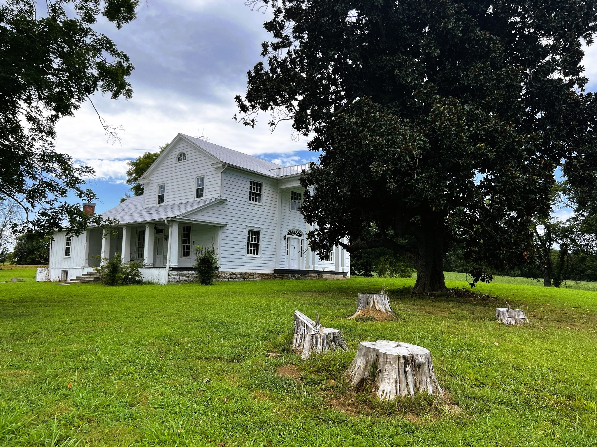 White two-story farmhouse with a covered porch, shaded by a large tree, with cut stumps across the lawn.